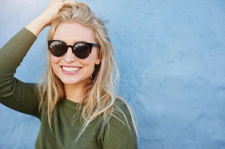 Smiling woman wearing sunglasses against a blue wall, showing healthy white teeth.